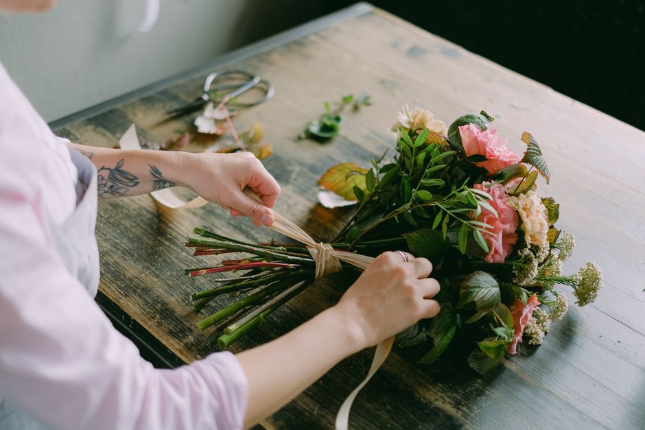 A person arranging a fresh floral bouquet on a wooden table, featuring pink and cream roses, white and green fillers, and assorted greenery. Their hands, with a visible tattoo on the forearm, are tyin