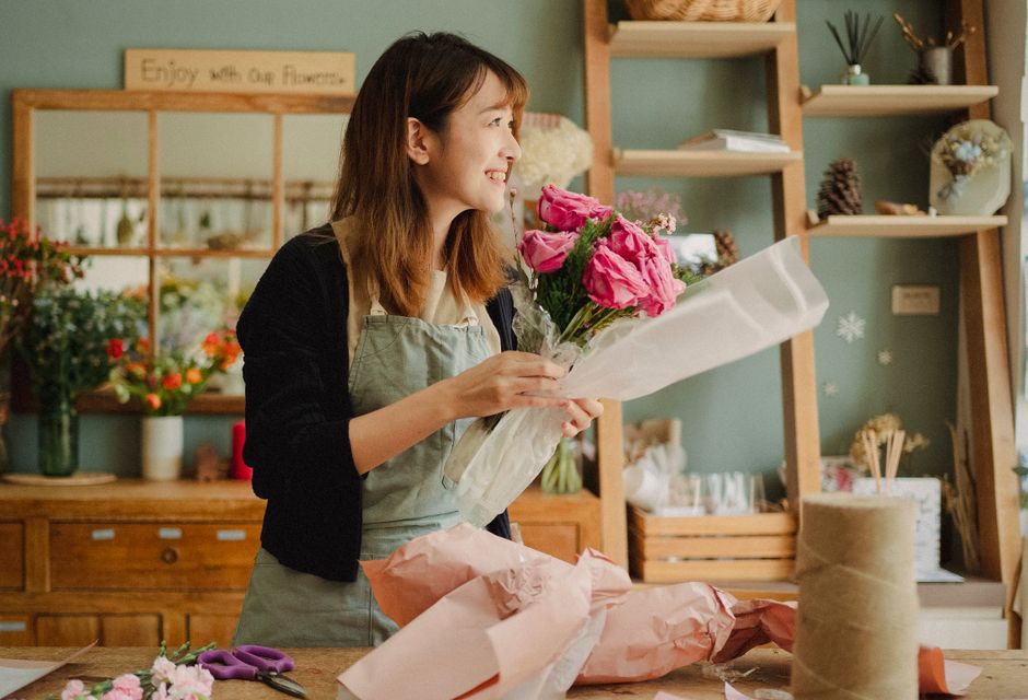 A smiling female florist with shoulder-length brown hair is holding a bouquet of vibrant pink roses and light purple flowers, wrapped in clear cellophane and white tissue paper, in a cozy flower shop 