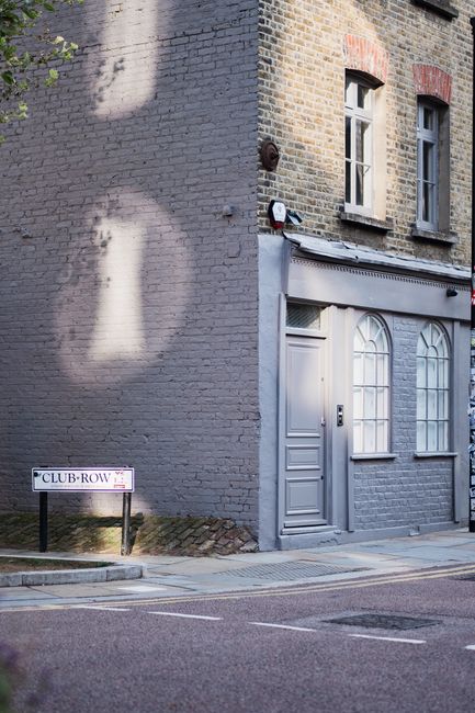 An exterior view of a charming brick building with pastel grey window frames and a matching door, located on a quiet street in Shoreditch. The building features two tall, arched windows with white tri