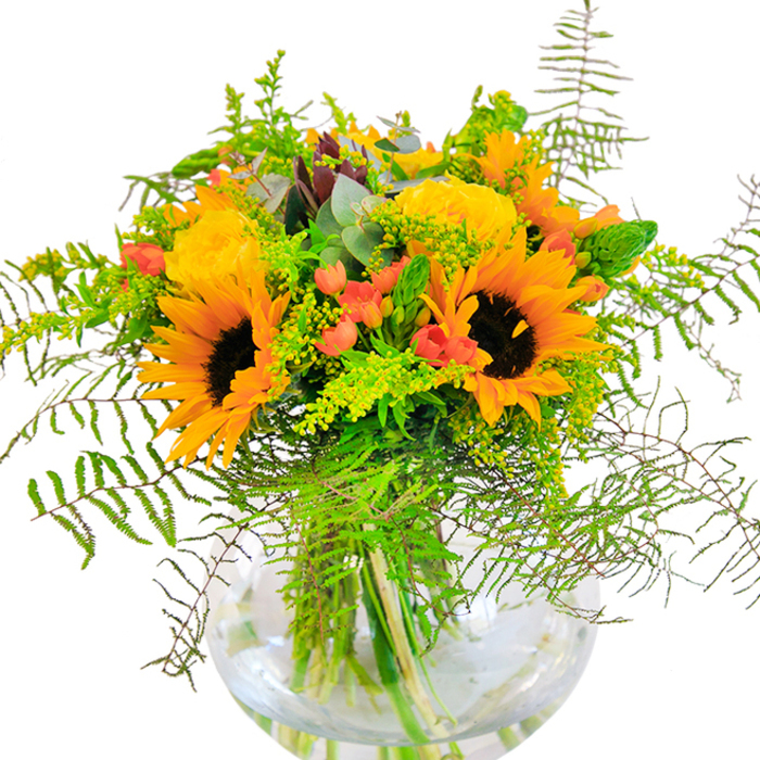 From the first glance, this abundant bouquet radiates the youthful vibrancy and artistic flair that defines Shoreditch. Sunflowers-iconic in their stature-dominate with dark centers circled by fiery yellow-orange petals, reminiscent of summer sunlight spilling over nearby rooftop terraces. Close by, tightly wound yellow garden roses spiral open in elegant succession, their plush texture amplifying a sense of comfort and sophistication. Scattered throughout are bursts of crimson-red hypericum berries, balancing the palette with invigorating energy that recalls the bold murals along Brick Lane. Feathery fern fronds extend outward, their soft textures and gentle cascades evoking the organic rhythm of local garden squares. Airy sprigs of goldenrod and slender shoots of green filler carry whispers of wild meadows, while subtle blue-green eucalyptus threads a modern edge into the mix. All of this floral artistry sits in a beautifully transparent, spherical glass vase-its water magnifying the interplay of stems, offering a view as inviting as any Shoreditch window display. The bouquet invites touch, with its contrast of silky rose petals, sturdy sunflowers, and crisp greenery. Light dances across the composition, suggesting optimism and celebration, making it ideal for birthdays, housewarmings, or transforming any East London living space into a gallery of joy and connection.